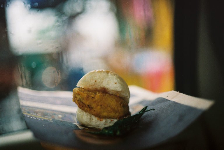 A vada pav on a plate at a street stall, the stall's colours blurred behind