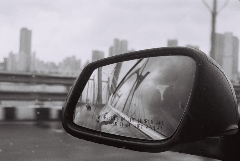 Black and white: a car's wing mirror reflecting the road, city towers beyond