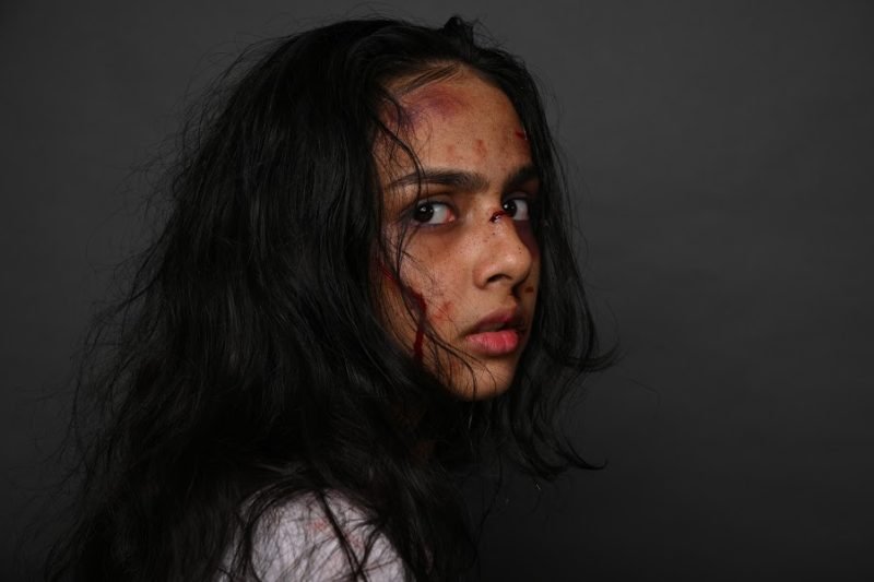 Special-effects makeup simulating cuts and bruising across the face, hair swept across, dark backdrop