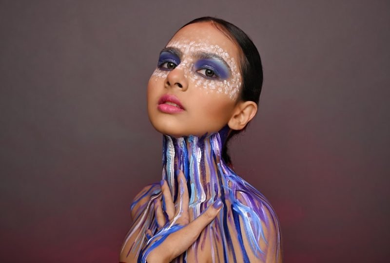 Editorial makeup: blue and purple paint running down the neck and chest, white dotted detail around the eyes