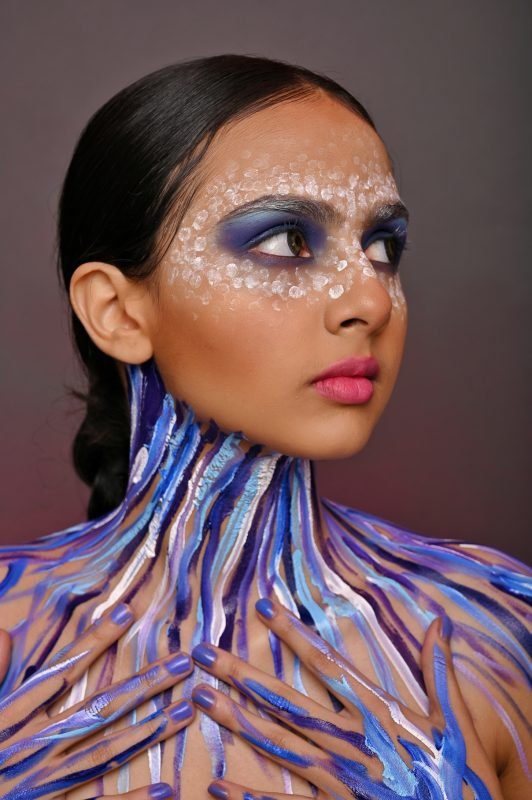 Editorial makeup in close-up: blue and purple paint streaking across the chest and throat, smoky blue eyes