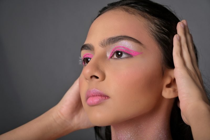 Graphic pink eye makeup with glitter on the cheekbones, hand raised beside the face