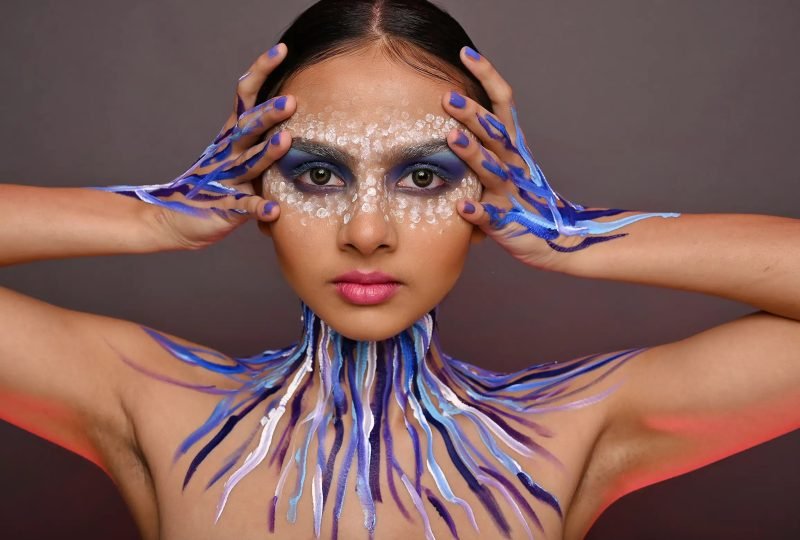 Editorial makeup: hands framing the face, blue and purple paint across the throat and shoulders