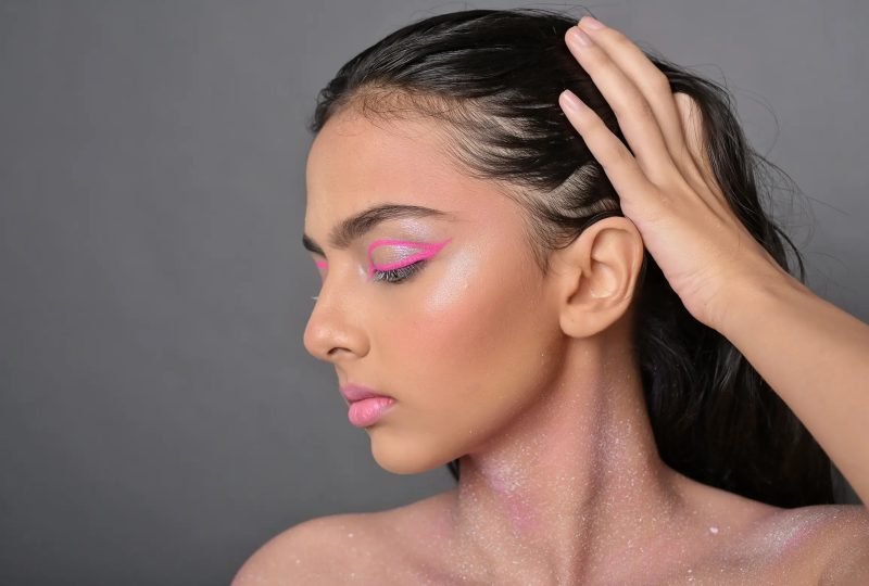 Profile portrait: pink eye makeup and glitter across the cheek, hand sweeping back the hair