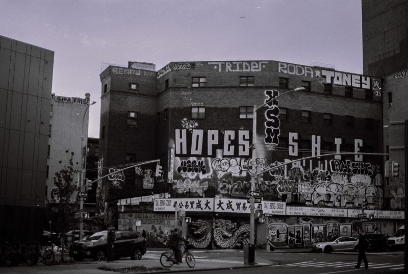 A street corner building covered in graffiti and posters, a cyclist passing below