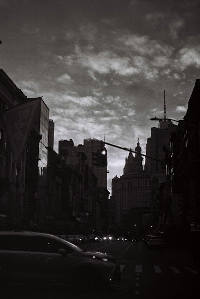 Black and white: a street at dusk, parked cars and a broken sky above the buildings