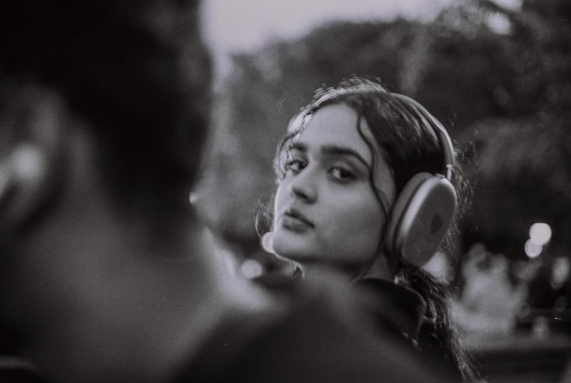 Black and white street portrait: a young woman in headphones glancing back over a passer-by's shoulder