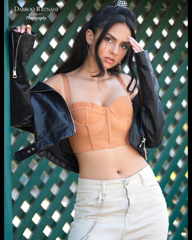 Sargam in an orange corset top with a black leather jacket off one shoulder, against a green lattice fence