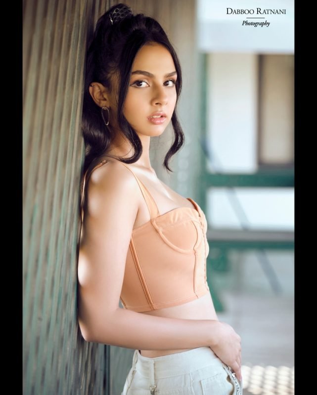 Sargam in an orange corset top and white trousers, turned to the side against a pale pillar