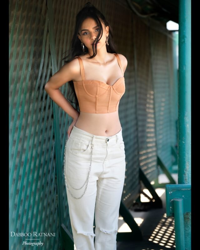 Sargam in an orange corset top and white jeans, hands raised to her hair in a green mesh corridor