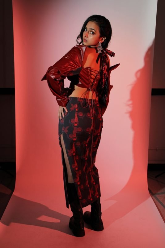 Rea by Rhea lookbook: a model with hands on hips in a red leather jacket and floral skirt, red backdrop