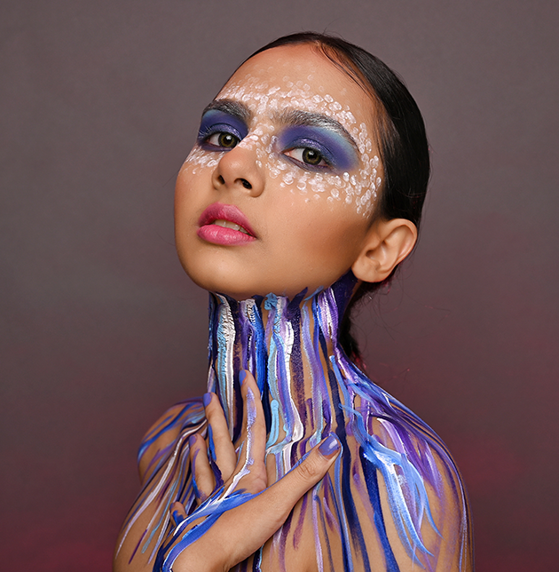 Editorial makeup: blue and purple paint running down the neck and chest, white dotted detail around the eyes