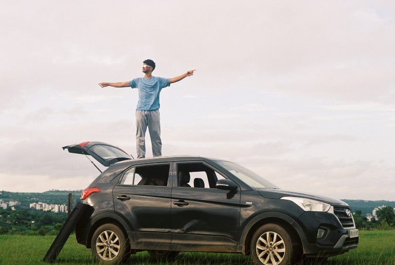 A person standing on the roof of a parked car with both arms outstretched, fields behind
